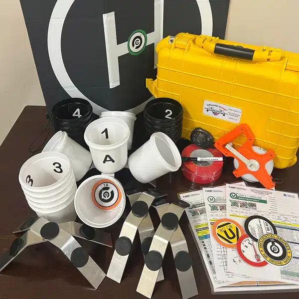 A tabletop collection of training and measuring gear: white cups labeled 1, 3, and 4, black cups labeled 2, a bright yellow hard plastic case, tape measures, orange hand tool, circular target stickers, and printed instruction sheets on a dark surface.